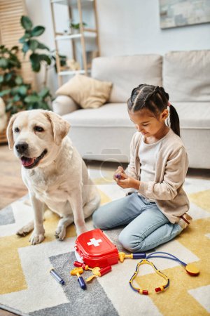 fröhliches Kind in Freizeitkleidung spielt Arzt mit Labrador im modernen Wohnzimmer, Spielzeug-Verbandskasten