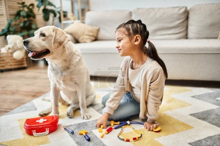 glückliches Mädchen in lässiger Kleidung spielt Arzt mit Labrador im modernen Wohnzimmer, Spielzeug-Verbandskasten
