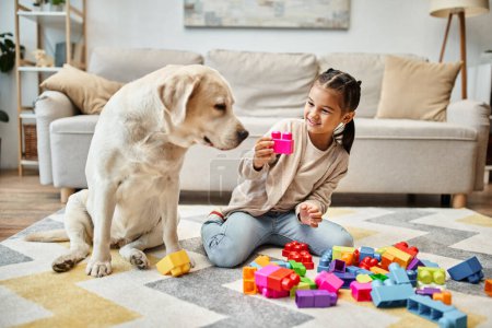 Foto de Chica feliz jugando con bloques de juguete de colores cerca de labrador en la sala de estar, torre de construcción juego - Imagen libre de derechos