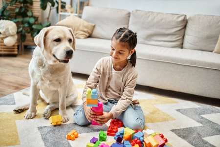 chica sonriente jugando con bloques de juguete de colores cerca de labrador en la sala de estar, torre de construcción juego