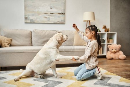 niedliches Mädchen in Freizeitkleidung, das mit Labrador spielt und im Wohnzimmer Leckereien gibt, Kind trainiert Hund
