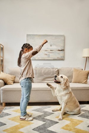 Mädchen in Freizeitkleidung trainiert Labrador in modernem Wohnzimmer, Kind gibt Leckerbissen beim Hundeunterricht