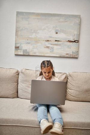 glückliches Mädchen im Grundschulalter, das auf dem Sofa sitzt und Laptop im modernen Wohnzimmer benutzt, E-Learning