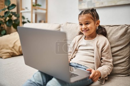 chica de edad elemental feliz sentado en el sofá y el uso de la computadora portátil en la sala de estar moderna, e-learning