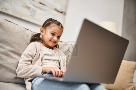Téléchargez les photos : Joyeuse fille d'âge élémentaire assis sur le canapé et en utilisant un ordinateur portable dans le salon moderne, e-learning - en image libre de droit