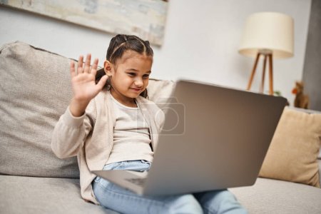 happy elementary age girl sitting on sofa and waving hand at laptop in living room, online lesson