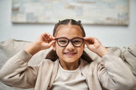 Téléchargez les photos : Heureuse fille d'âge élémentaire en tenue décontractée portant des lunettes et regardant la caméra dans le salon - en image libre de droit