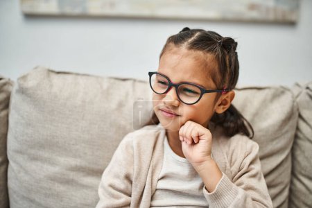 Nachdenkliches Mädchen im Grundschulalter in lässiger Kleidung mit Brille und wegschauendem Blick im Wohnzimmer