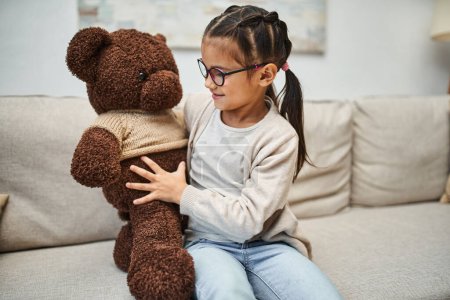 glückliches Mädchen in Freizeitkleidung und Brille umarmt weichen Teddybär und sitzt auf dem Sofa im Wohnzimmer
