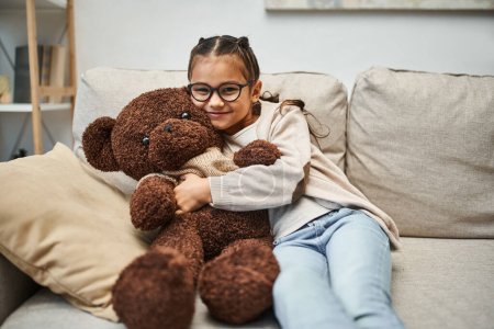 Foto de Chica feliz en ropa casual y anteojos abrazando oso de peluche suave y sentado en el sofá en la sala de estar - Imagen libre de derechos