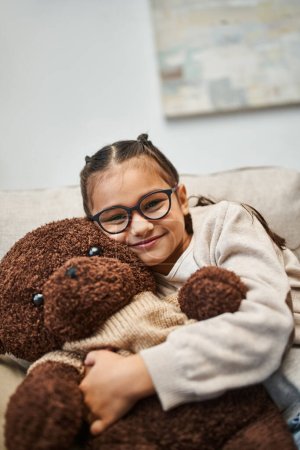 fröhliches Mädchen in Freizeitkleidung und Brille umarmt weichen Teddybär und sitzt auf dem Sofa im Wohnzimmer