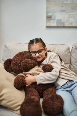 happy kid in casual wear and eyeglasses hugging soft teddy bear and sitting on sofa in living room