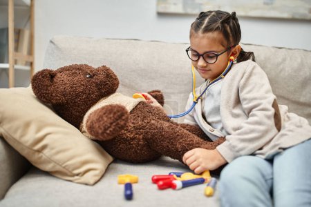 happy kid in casual wear and eyeglasses playing doctor with teddy bear on sofa in living room