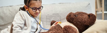 happy kid in casual wear and eyeglasses playing doctor with teddy bear on sofa in living room