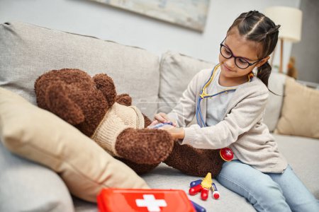 Téléchargez les photos : Enfant heureux en tenue décontractée et lunettes jouant médecin avec ours en peluche sur canapé dans le salon - en image libre de droit