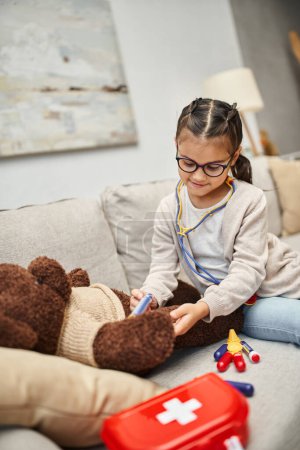 happy child in casual wear and eyeglasses playing doctor with teddy bear on sofa in living room