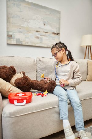 glückliches Kind in Freizeitkleidung und Brille spielt Arzt mit weichem Teddybär auf Sofa im Wohnzimmer