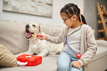 glückliches Kind in Freizeitkleidung und Brille spielt Arzt mit Labrador-Hund auf Sofa im Wohnzimmer