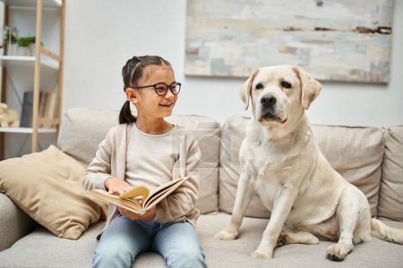 fröhliches Mädchen in Freizeitkleidung und Brille liest Buch neben Labrador-Hund auf Sofa im Wohnzimmer
