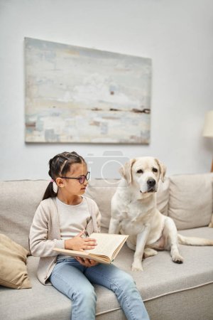 cute girl in casual wear and eyeglasses reading book near labrador dog on sofa in living room