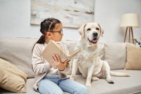 Téléchargez les photos : Fille intelligente en tenue décontractée et lunettes de lecture livre pour chien labrador sur canapé dans le salon - en image libre de droit