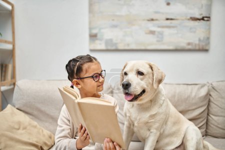 Téléchargez les photos : Mignonne fille en tenue décontractée et lunettes de lecture livre près de chien labrador sur canapé dans le salon - en image libre de droit