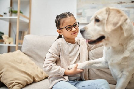 glückliches Mädchen im Grundalter mit Brille, die Pfote eines Labradors haltend und auf dem Sofa im Wohnzimmer sitzend