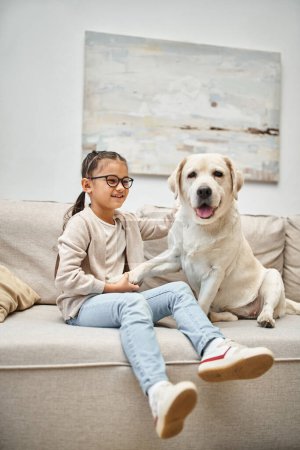 glückliches Mädchen im Grundalter mit Brille, die Pfote eines Labradors haltend und auf dem Sofa im Wohnzimmer sitzend