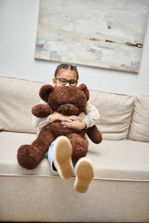 entzückendes Mädchen im Grundschulalter mit Brille und Teddybär auf dem Sofa im Wohnzimmer