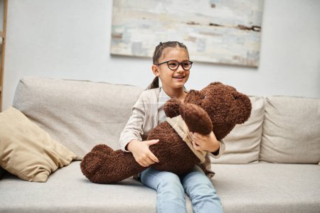 adorable elementary age kid in eyeglasses holding teddy bear and sitting on sofa in living room