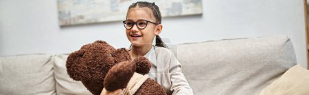 adorable elementary age girl in eyeglasses holding teddy bear and sitting on sofa, banner