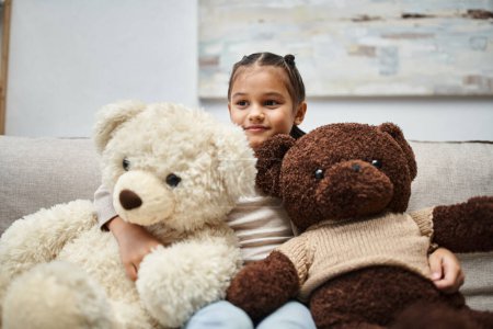 Niedliche Grundschulkind in Freizeitkleidung sitzt auf dem Sofa mit weichen Teddybären im Wohnzimmer