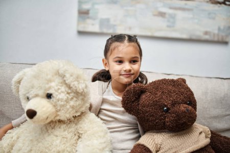 Nettes Mädchen im Grundschulalter in Freizeitkleidung sitzt auf dem Sofa mit weichen Teddybären im Wohnzimmer