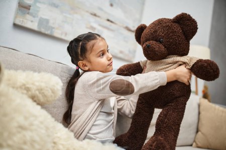 Niedliche Grundschulkind in Freizeitkleidung sitzt auf dem Sofa mit weichen Teddybären im Wohnzimmer