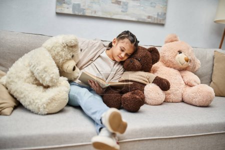 Photo for Adorable girl sitting on sofa with soft teddy bears and reading book in modern living room - Royalty Free Image