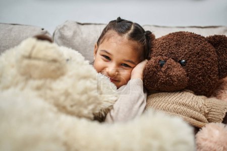fröhliches Mädchen im Grundalter sitzt zwischen weichen Teddybären auf der Couch im modernen Wohnzimmer