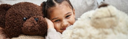 Photo for Cheerful elementary age girl sitting among soft teddy bears on couch in modern living room, banner - Royalty Free Image