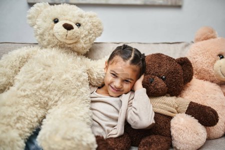 Photo for Cheerful elementary age girl sitting among soft teddy bears on couch in modern living room, at home - Royalty Free Image
