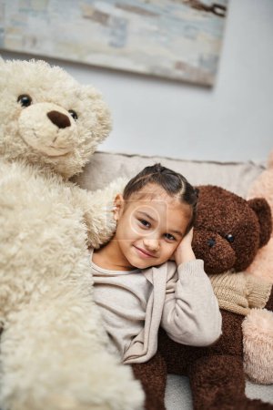 Nettes Mädchen im Grundschulalter sitzt zwischen weichen Teddybären auf der Couch im modernen Wohnzimmer