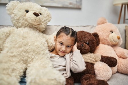 entzückendes Mädchen im Grundschulalter sitzt zwischen weichen Teddybären auf der Couch im modernen Wohnzimmer