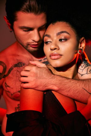 guapo hombre tatuado abrazando amorosamente a su novia afroamericana por detrás, sexy pareja