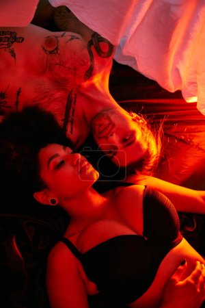 Téléchargez les photos : Attrayant couple multiculturel couché sensuellement ensemble sous drap de lit entouré de lumières - en image libre de droit