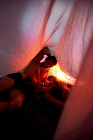 sexy multikulturelles Paar in Unterwäsche liegt sinnlich zusammen unter Bettlaken, umgeben von Lichtern
