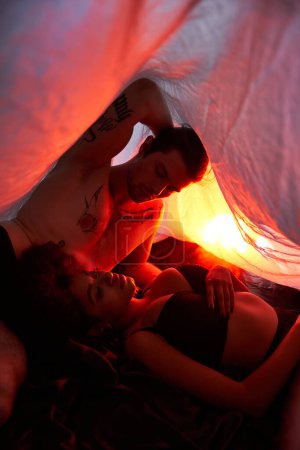 diverse young couple in underwear lying sensually together under bedsheet surrounded by lights