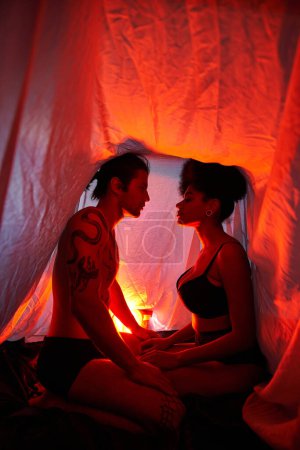 Téléchargez les photos : Couple multiculturel sensuel assis sous le drap de lit et posant amoureusement entouré de lumières - en image libre de droit