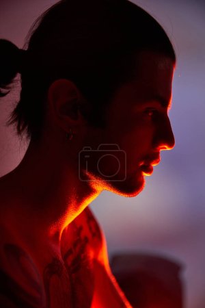 joven hombre guapo con tatuajes en el cuerpo y pendientes posando en perfil rodeado de vibrantes luces