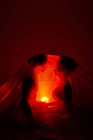 Multikulturelles Paar mit Bettlaken bedeckt, umgeben von Licht auf dunklem Hintergrund, Zweisamkeit