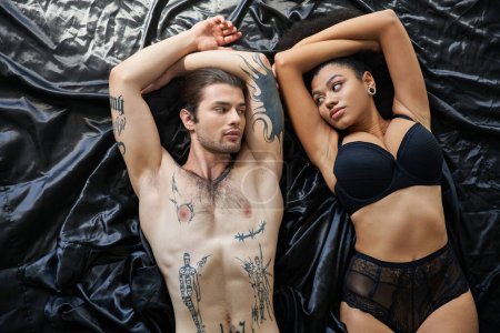 Téléchargez les photos : Jeune femme afro-américaine en lingerie couchée au lit à côté de son petit ami tatoué, couple sexy - en image libre de droit