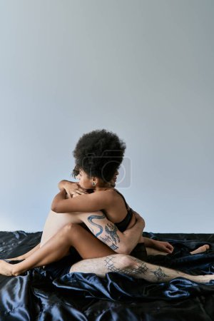 Téléchargez les photos : Sexy jeune couple multiculturel assis sur leur lit et câlin avec amour sur fond gris - en image libre de droit