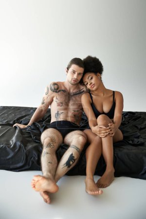 Téléchargez les photos : Interracial beau couple en sous-vêtements assis près de l'autre sur le lit, ensemble - en image libre de droit